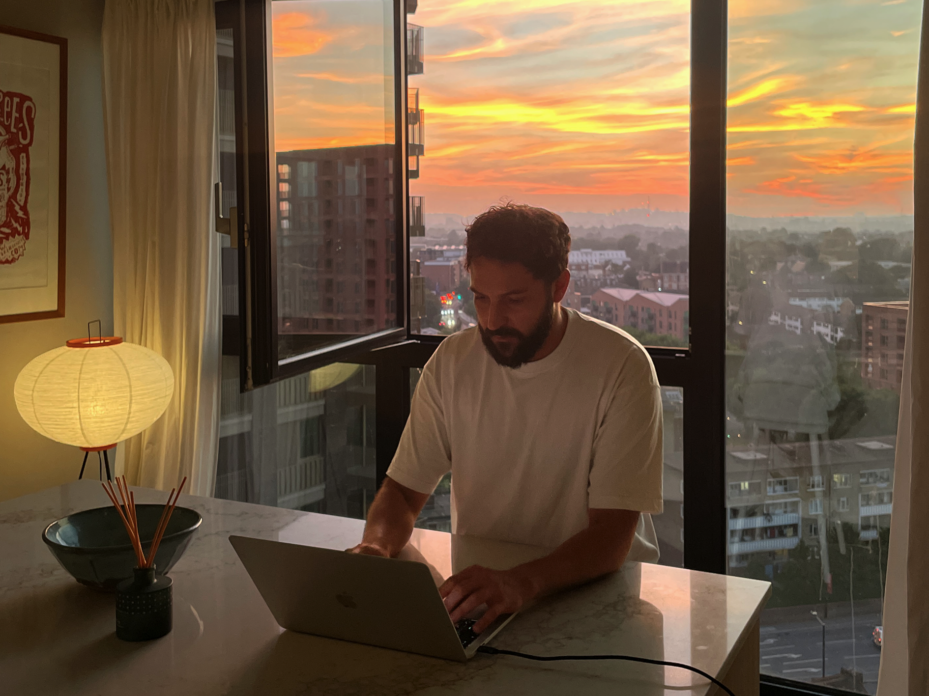 Man working on a laptop in a room with a large window view of a sunset over a cityscape.